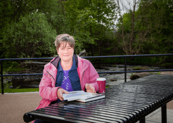 Tracey McLennan wears a pink jacket and sits with a coffee and a pile of books next to a river.